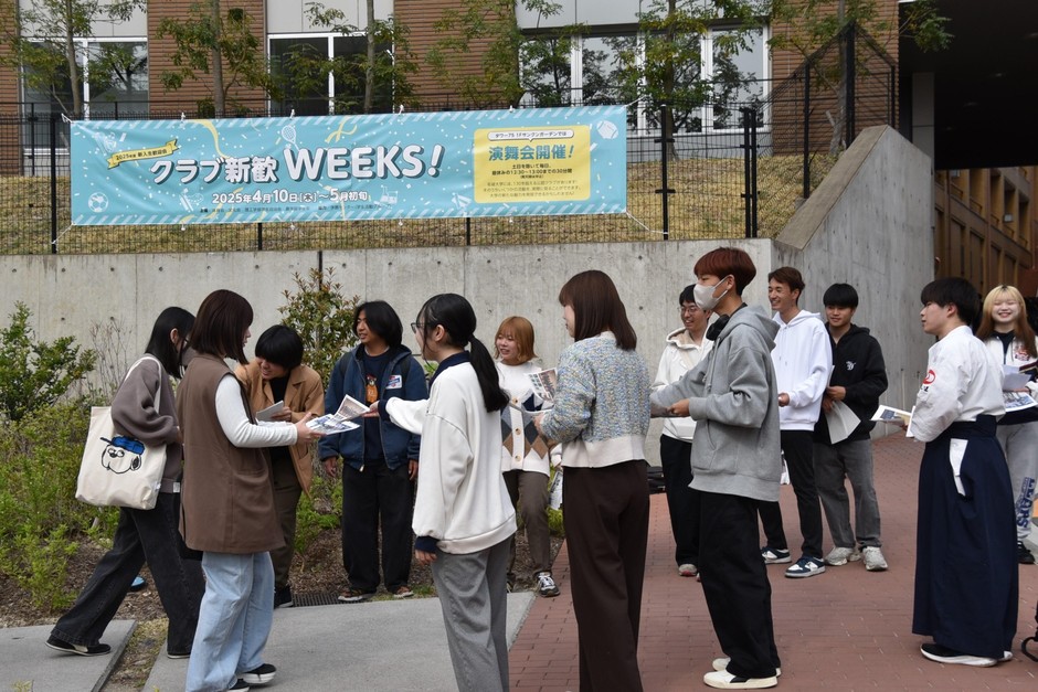 「クラブ新歓WEEKS!」の告知の前でビラ配り